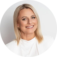 Portrait of a smiling blonde woman in a white top against a light gray background, circular crop.